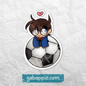 May include: A sticker featuring a cartoon character with brown hair, glasses, and a blue jacket, perched on a soccer ball. A pink heart floats above the character's head. The sticker has the website gabapple.com printed on it.