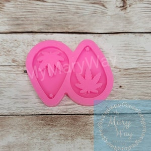 May include: Pink silicone mold for making earrings. The mold has two cavities, one with a marijuana leaf design and the other with a palm tree design.