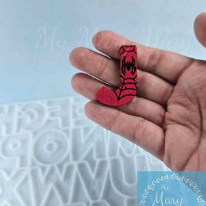 May include: Red wooden letter J with a black spider web design and a black spider in the center. The letter is being held in a hand.