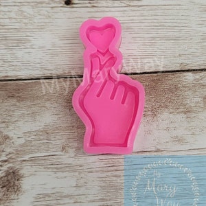 May include: Pink silicone mold of a hand making a heart shape with a finger. The mold is on a wooden surface.