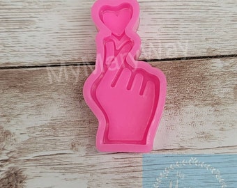May include: Pink silicone mold of a hand making a heart shape with a finger. The mold is on a wooden surface.