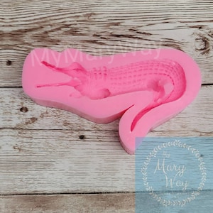 May include: Pink silicone mold of an alligator with a textured back. The mold is on a wooden surface.