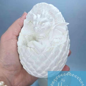 May include: A white dragon sculpture emerging from a textured egg. The dragon has detailed wings and scales. The egg is held in a hand, with a blue background. The image includes the text "My Mary Way Mixed Media Art".