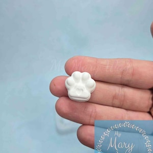 May include: White ceramic paw print charm with a small heart in the center. The charm is being held in a hand.