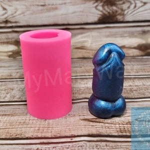 May include: A bright pink silicone mold and a blue and purple resin sculpture. The mold is cylindrical with a rounded top. The sculpture is a stylized representation of a male anatomy.