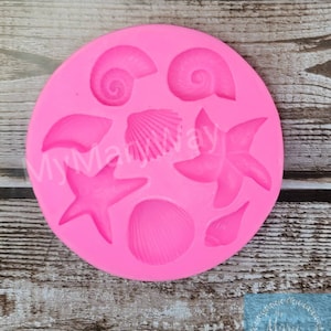 May include: Pink silicone mold with seven different shapes of seashells and starfish. The mold is on a wooden surface.
