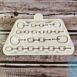 May include: A white silicone mold with multiple shapes of chains and rings. The mold is designed for making fondant or gum paste decorations. The mold is labeled "©2019 Katy Sua Designs LTD".