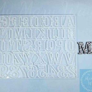May include: White plastic mold with letters A through Z and numbers 0 through 9. The mold is on a white surface. A black and white letter M is to the right of the mold.