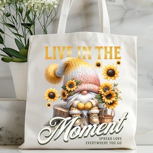 Może przedstawiać: Biała torba typu tote z żółtym gnomem trzymającym słonecznik. Na torbie nadrukowany jest tekst "Live in the Moment" i "Spread Love Everywhere You Go".