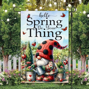 May include: A colourful garden flag featuring a gnome wearing a red and black hat and holding a watering can. The flag reads "hello spring do your thing" with butterflies and ladybirds.