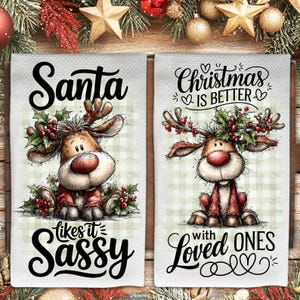 Puede incluir: Dos paños de cocina navideños blancos con ilustraciones de renos. Un paño dice "Santa likes it Sassy" y el otro "Christmas is better with Loved Ones". Los renos tienen narices rojas y visten trajes rojos.