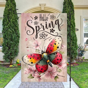May include: A garden flag featuring a colorful butterfly with red, yellow, and green wings, and black spots. The flag has the words "Hello Spring" in a decorative font, with floral accents. The background is a soft pink.