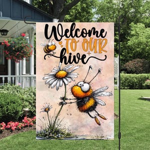 May include: A garden flag with the words "Welcome to our hive" in black and orange. The flag features cartoon bees and daisies. The background is a light pink color.