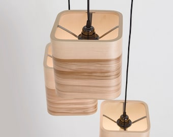 Könnte beinhalten: Drei moderne Pendelleuchten mit quadratischen Holzschirmen. Die Schirme bestehen aus hellem Holz mit dunkleren horizontalen Streifen. Jede Leuchte hängt an einem schwarzen Kabel, und das Innere der Schirme leuchtet mit warmem Licht.