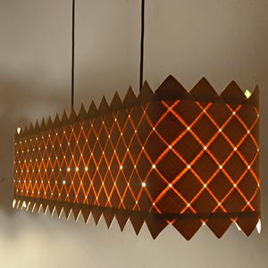 May include: A rectangular pendant light with a diamond pattern and a zig-zag top edge. The light emits a warm orange glow, and the fixture is suspended from the ceiling by two black cords.
