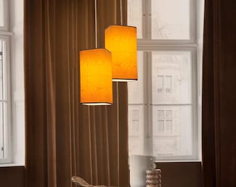 Könnte beinhalten: Zwei zylindrische Pendelleuchten mit orangefarbenen Stoffschirmen hängen von der Decke. Die Schirme haben ein strukturiertes Muster und eine dunkle Umrandung. Der Hintergrund umfasst braune Vorhänge und einen Tisch mit Dekorationsartikeln.
