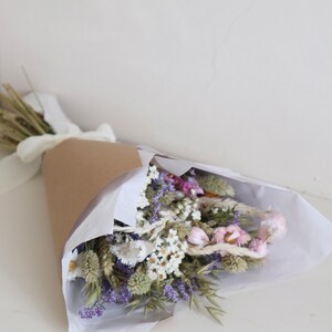 May include: A bouquet of dried flowers wrapped in brown paper and white tissue. The bouquet features a variety of flowers in shades of purple, white, and pink, tied with a white ribbon. The arrangement is presented on a white surface.