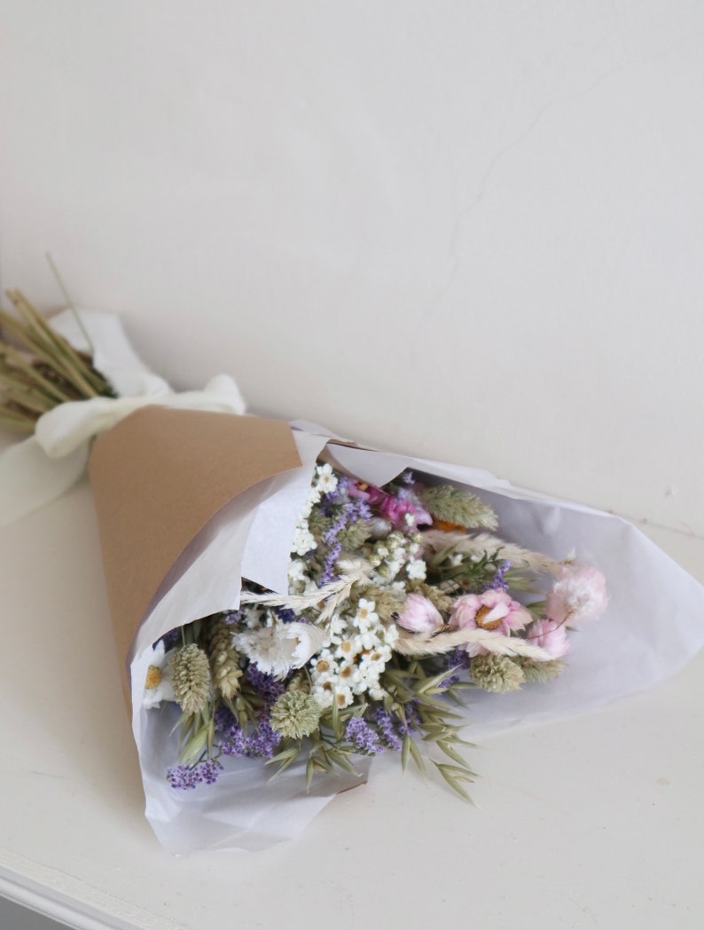 May include: A bouquet of dried flowers wrapped in brown paper and white tissue. The bouquet features a variety of flowers in shades of purple, white, and pink, tied with a white ribbon. The arrangement is presented on a white surface.