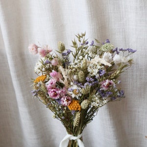 May include: A dried flower bouquet featuring a variety of colors, including pink, purple, yellow, and white. The bouquet is tied with a white ribbon. The flowers are arranged against a neutral background.