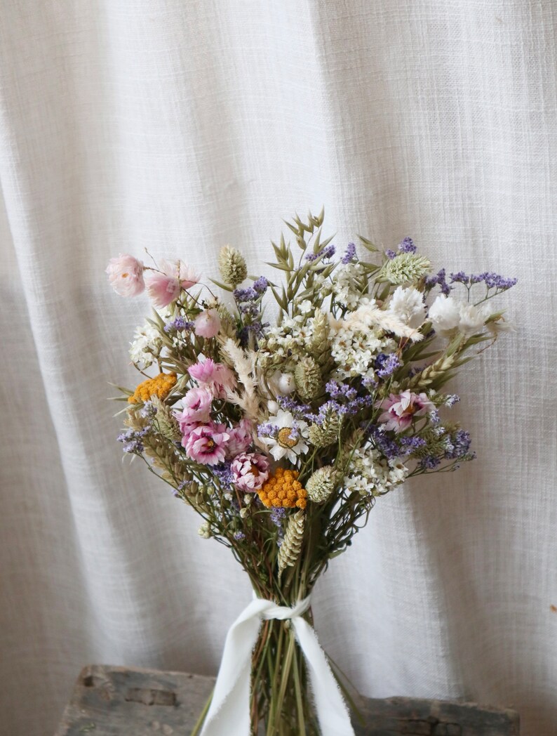 May include: A dried flower bouquet featuring a variety of colors, including pink, purple, yellow, and white. The bouquet is tied with a white ribbon. The flowers are arranged against a neutral background.