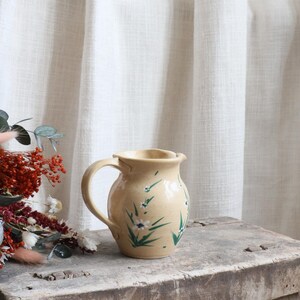 May include: A cream-coloured ceramic jug with a handle, decorated with hand-painted white flowers and green leaves. The jug is placed on a rustic wooden surface, with dried flowers in the background. A charming piece of kitchenware.
