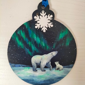 May include: A round wooden ornament painted with a dark blue night sky, green and blue aurora borealis, and two white polar bears. A white snowflake is attached to the top of the ornament.