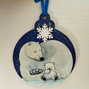 May include: A blue and white wooden ornament with a painted image of a polar bear and a cub. The ornament is decorated with a white snowflake and a blue ribbon.