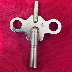 May include: A silver-colored watch key with a cylindrical handle and a keyhole-shaped head. The head has two circular openings and the numbers "6" and "17" are visible. The key is set against a red background.
