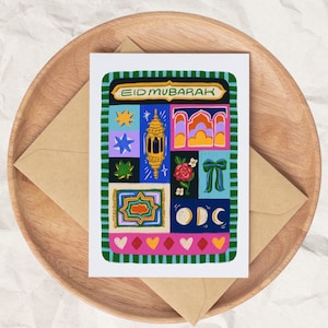 May include: A greeting card with the words "EID MUBARAK" in a gold banner. The card features colorful illustrations of a star, a lantern, an arch, a rose, a bow, a moon, and a rug. The card is on a wooden tray with a brown envelope.
