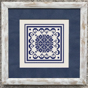May include: A framed cross-stitch artwork with a symmetrical blue and white geometric pattern. The artwork is set against a navy blue mount and framed in a distressed white wooden frame. The piece is displayed against a textured grey wall.