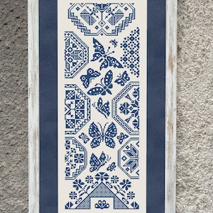 May include: A framed cross-stitch embroidery featuring a blue and white design of butterflies and geometric patterns. The embroidery is framed in a white wood frame with a blue mount.