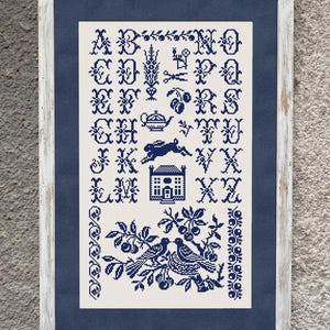 May include: A framed cross-stitch sampler featuring a dark blue alphabet on a cream background. The design includes decorative motifs such as a rabbit, a house, a teapot, scissors, and two birds perched on a branch, all enclosed in a weathered white wooden frame.
