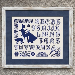 May include: Cross-stitch sampler in a white wooden frame. The design features a woman seated at a table with a cat, surrounded by an alphabet and decorative borders, all in navy blue on an off-white background.