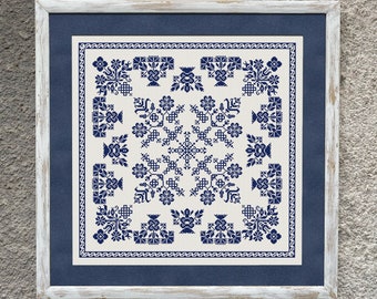Op de afbeelding: Een ingelijst borduurwerk met kruissteek in een blauw en wit geometrisch patroon. Het ontwerp heeft een centraal vierkant met een herhalend patroon van gestileerde bloemen en bladeren.