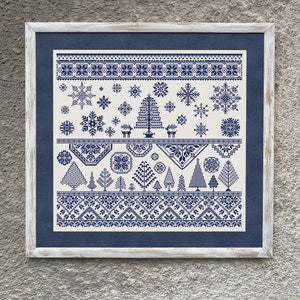 Op de afbeelding: Een wit ingelijst borduurwerk met een blauw en wit winters tafereel met sneeuwvlokken, bomen en geometrische patronen.