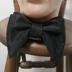 May include: A black bow tie made of a textured fabric. The bow tie is tied and displayed on a wooden mannequin.
