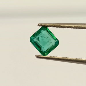Puede incluir: Una esmeralda verde vibrante, tallada en forma cuadrada, se sostiene con unas pinzas sobre un fondo blanco.