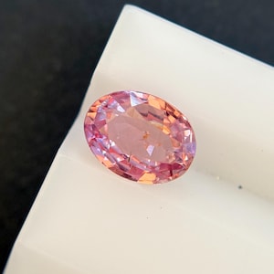May include: An oval-cut gemstone with a faceted surface displaying a gradient of pink and orange hues. The stone is translucent and reflects light across its polished surface, showcasing a vibrant color transition from soft pink to warm orange.