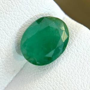 Puede incluir: Una gema de esmeralda verde vibrante, tallada en forma ovalada, se muestra sobre una superficie blanca.
