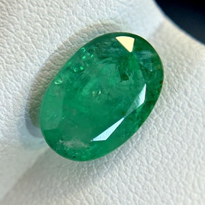 Puede incluir: Una esmeralda ovalada de color verde vibrante con un corte facetado. La esmeralda está colocada sobre un fondo blanco.