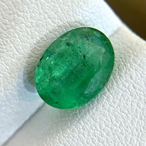 Puede incluir: Una esmeralda ovalada de color verde vibrante con inclusiones, sobre un fondo blanco.