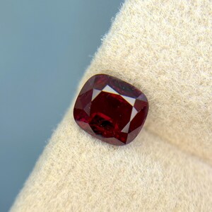 May include: A deep red, cushion-cut gemstone with light-reflecting facets. The gemstone is presented on a cream-coloured fabric, against a light blue backdrop. The gemstone's colour is a rich, saturated red.