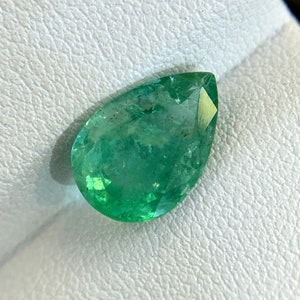 Puede incluir: Una piedra preciosa de esmeralda en forma de pera con un tono verde y un ligero tono amarillo. La piedra preciosa está colocada sobre un fondo blanco.