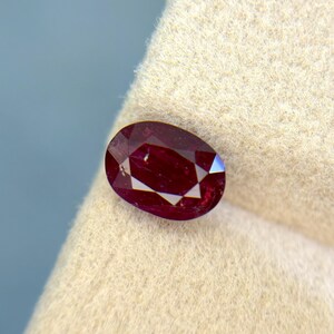 Könnte beinhalten: Ein facettierter, oval geformter Rubin-Edelstein. Der Rubin hat eine tiefe, satte rote Farbe mit sichtbaren inneren Facetten. Der Edelstein liegt vor einem neutralen Hintergrund, der seine Brillanz und Farbe hervorhebt. Eine Nahaufnahme zeigt die Klarheit des Edelsteins.