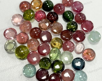 May include: A collection of round, multi-colored gemstones. The gemstones are various shades of pink, green, yellow, and blue. The gemstones are arranged in a pile on a white surface.