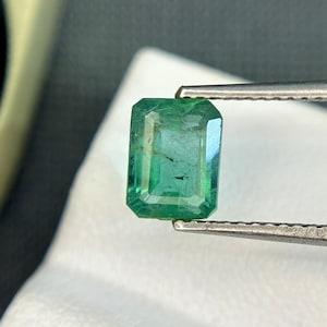 Puede incluir: Una gema de esmeralda verde vibrante, tallada en forma de esmeralda, se sujeta con unas pinzas. La gema es transparente y tiene una ligera inclusión.