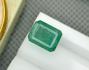 May include: A rectangular, emerald-cut green gemstone with a white border. The gemstone is set on a white surface, with a gold-colored object in the background. The gemstone has a vibrant green color and a clear, polished surface.