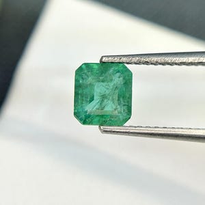 Puede incluir: Una esmeralda verde vibrante, tallada en forma cuadrada, se sostiene con unas pinzas. La esmeralda tiene una apariencia ligeramente nublada, lo que indica su origen natural.