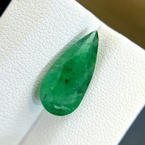 Puede incluir: Una piedra preciosa de esmeralda en forma de pera con un color verde intenso. La piedra preciosa tiene un corte facetado y está colocada sobre un fondo blanco.