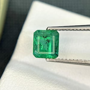 Puede incluir: Una gema de esmeralda verde vibrante con un corte cuadrado, sostenida por unas pinzas. La esmeralda tiene una ligera inclusión, visible como una mancha oscura.